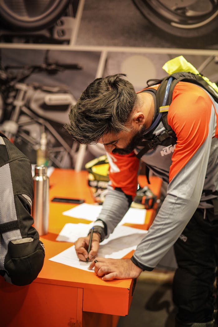 A motorcyclist in gear signs documents at an indoor workshop table.